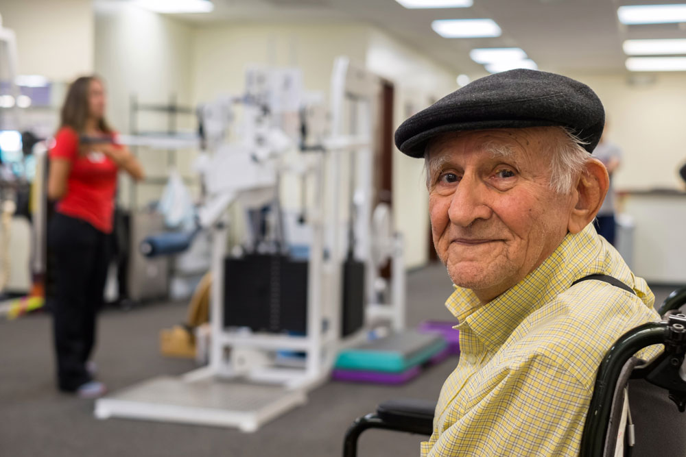 Elderly man sitting on a chair smiling with a woman behind doing arthritis exercises