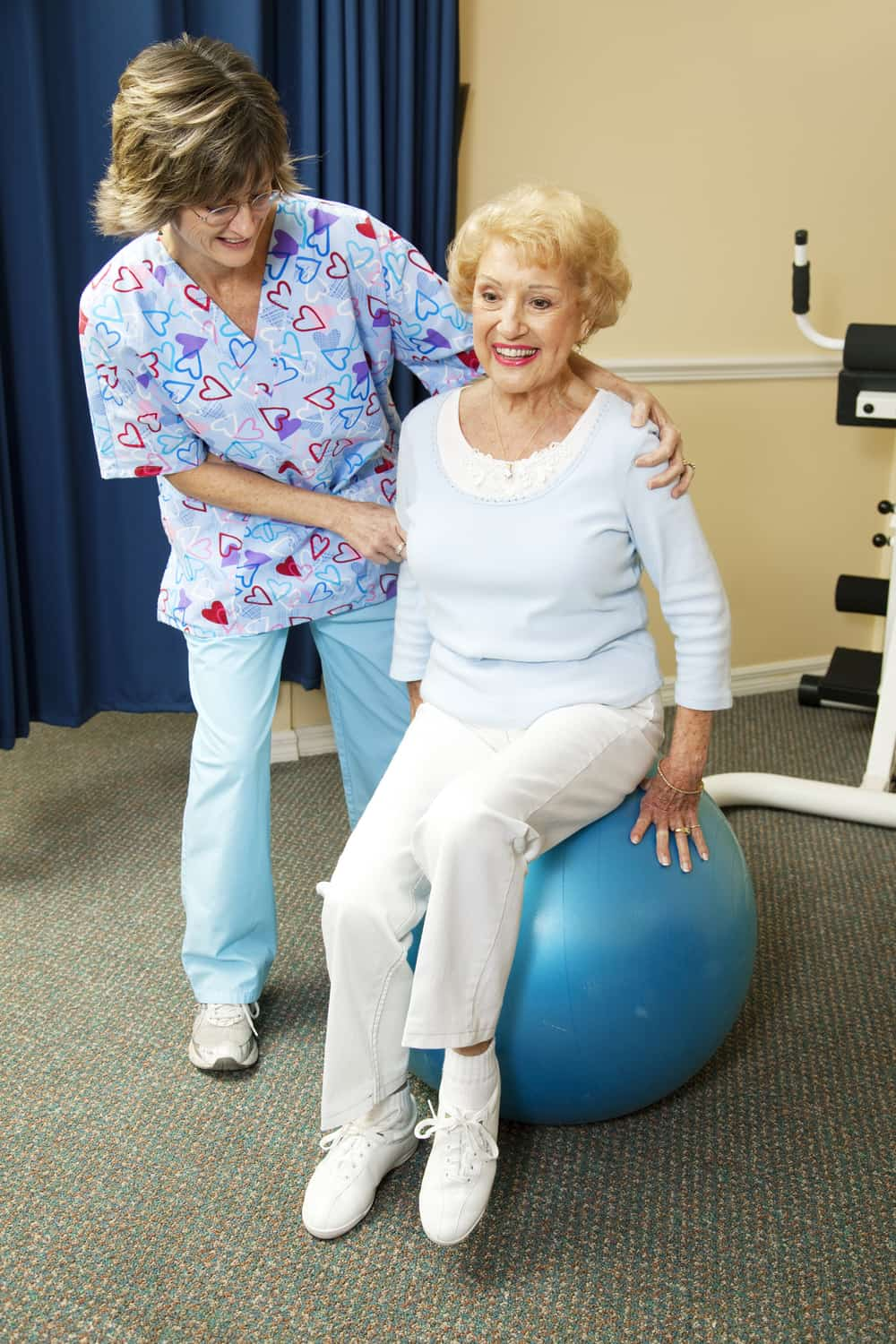 Nurse helping elderly woman doing physical therapy exercise using gym ball