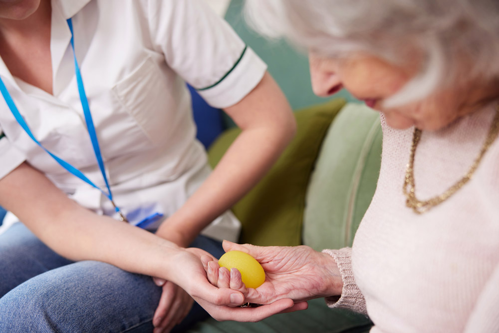 Female Physiotherapist Getting Senior Woman To Squeeze Rubber Ball At Home as part of occupational therapy for arthritis