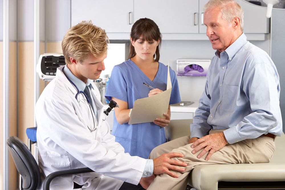 Doctor examining a senior male patient who had knee surgery and going through therapy while the nurse is taking notes.