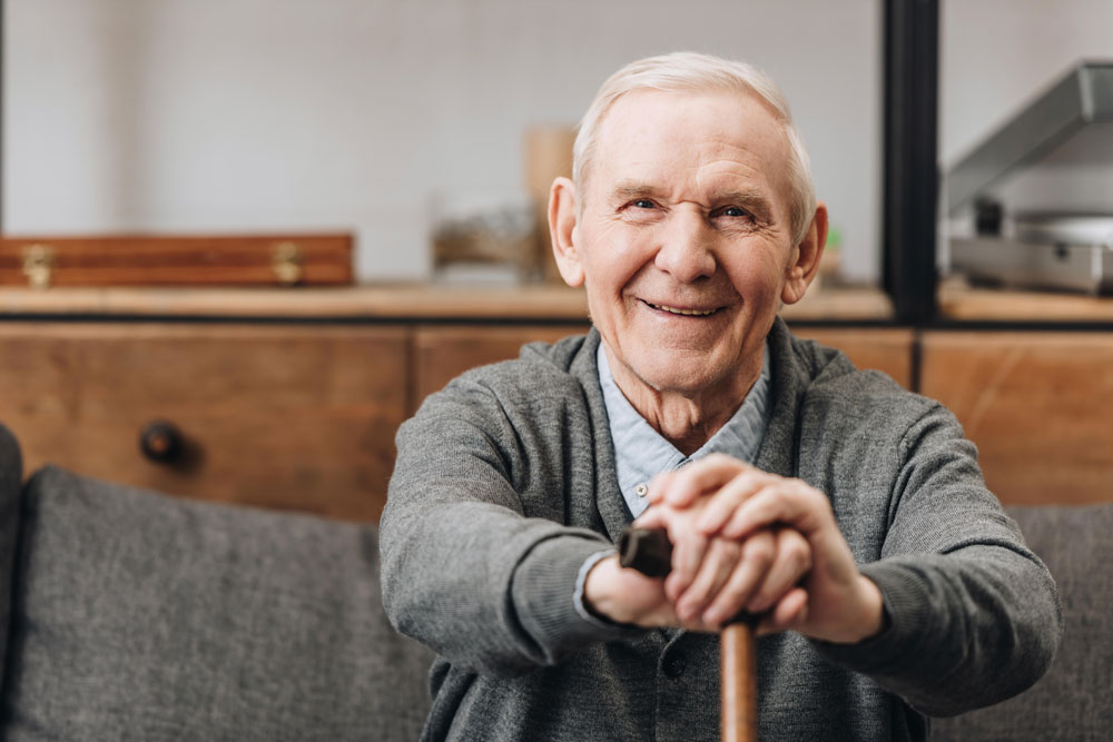 Cheerful old man suffering having common first signs of parkinson’s disease sitting on sofa smiling and holding walking cane