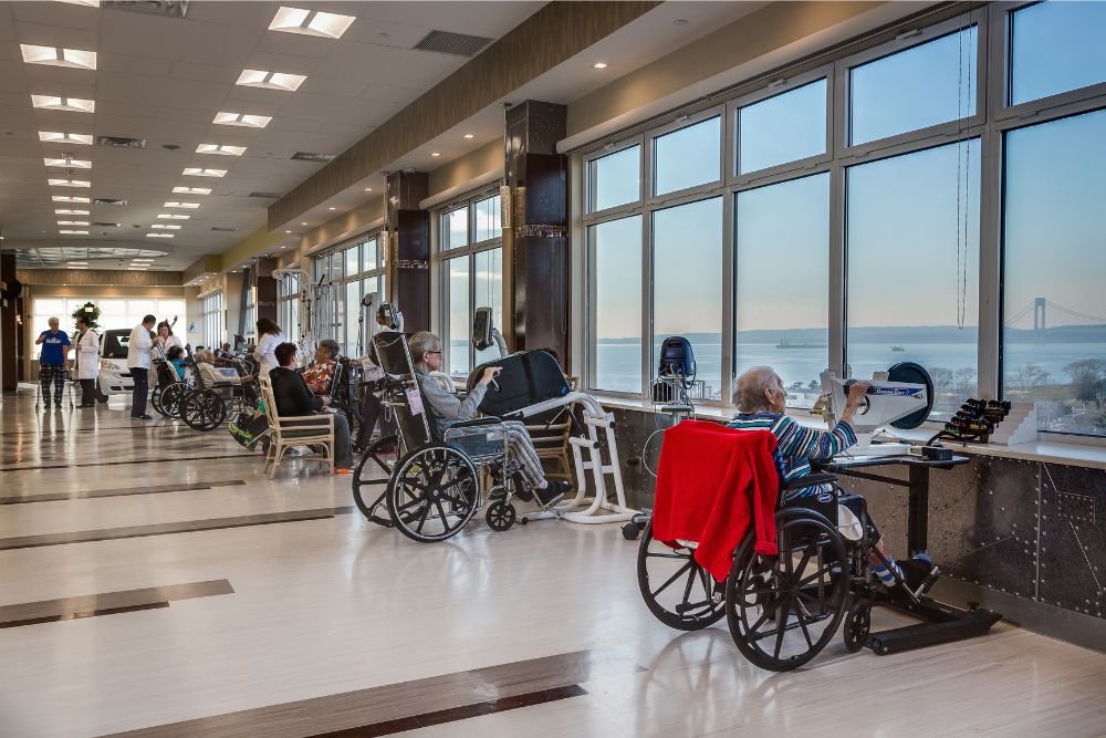 Elderly people doing physical therapy while enjoying the beautiful view of New York from the big window at Haym Salomon Home which provides the best aftercare for stents in the heart.