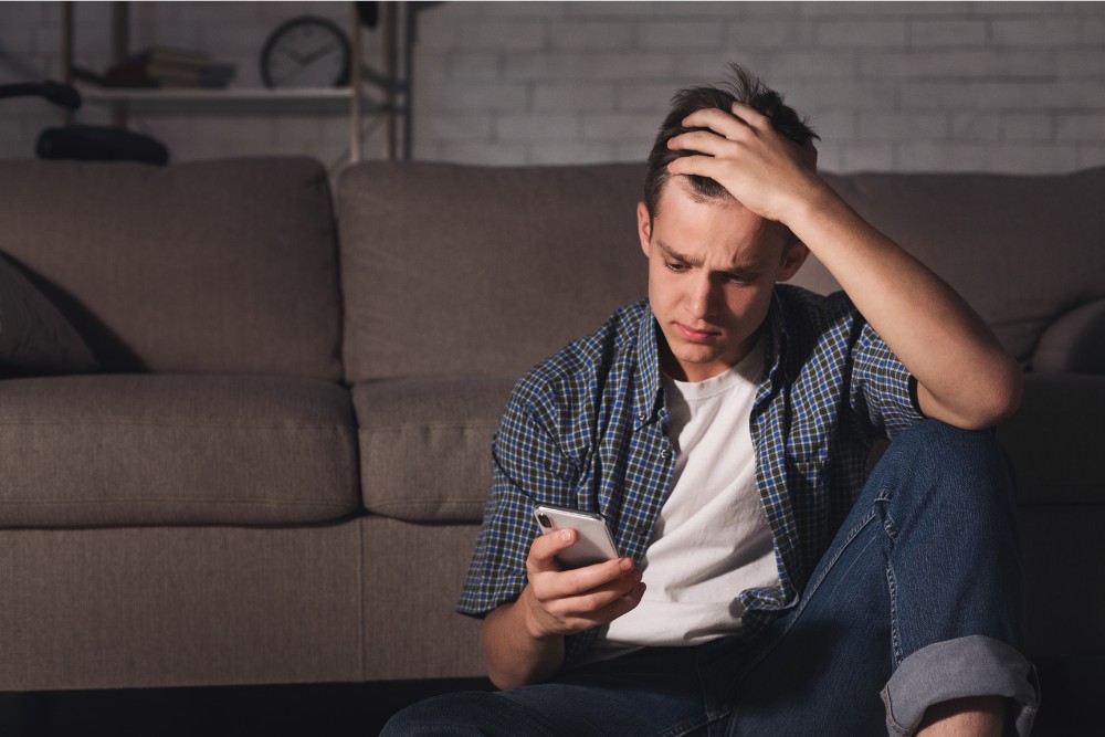Young man sitting on the floor with a distressed expression while looking at his phone, illustrating screen overuse and other brain damage habits.